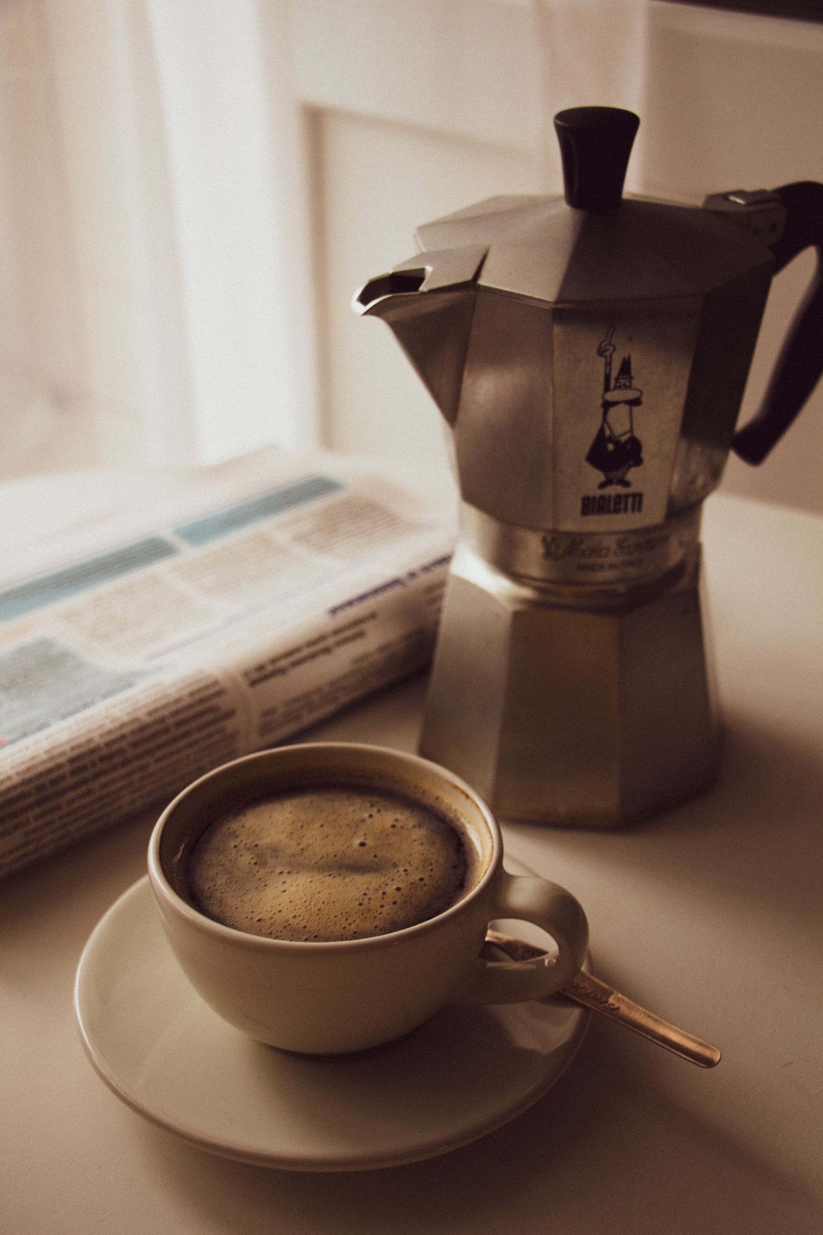 A warm cup of coffee next to a moka pot and a newspaper in a cozy morning setting