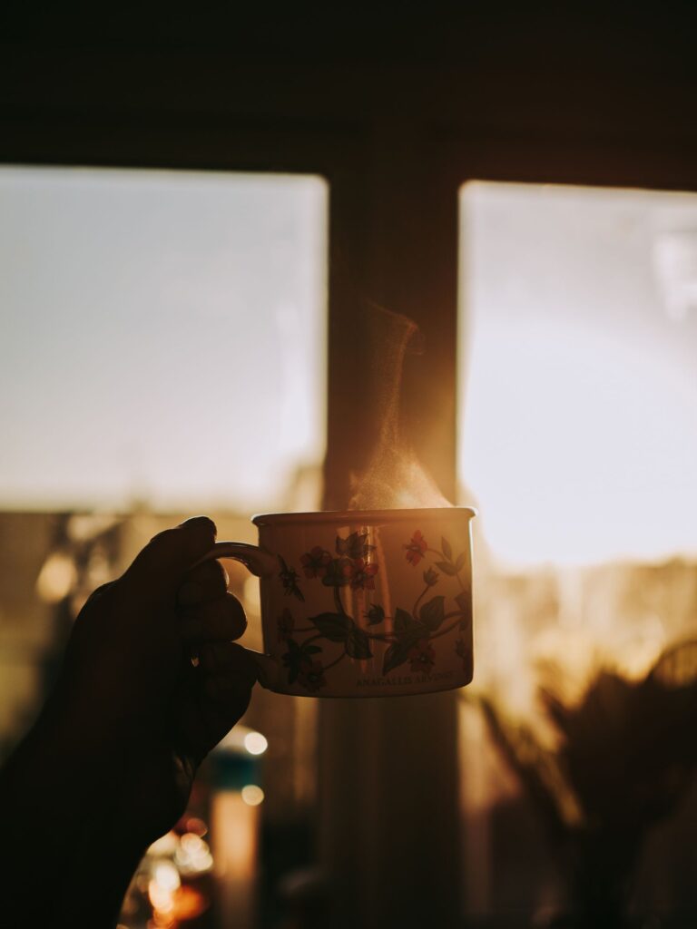 A hand holding a coffee mug against a bright sunrise through a window