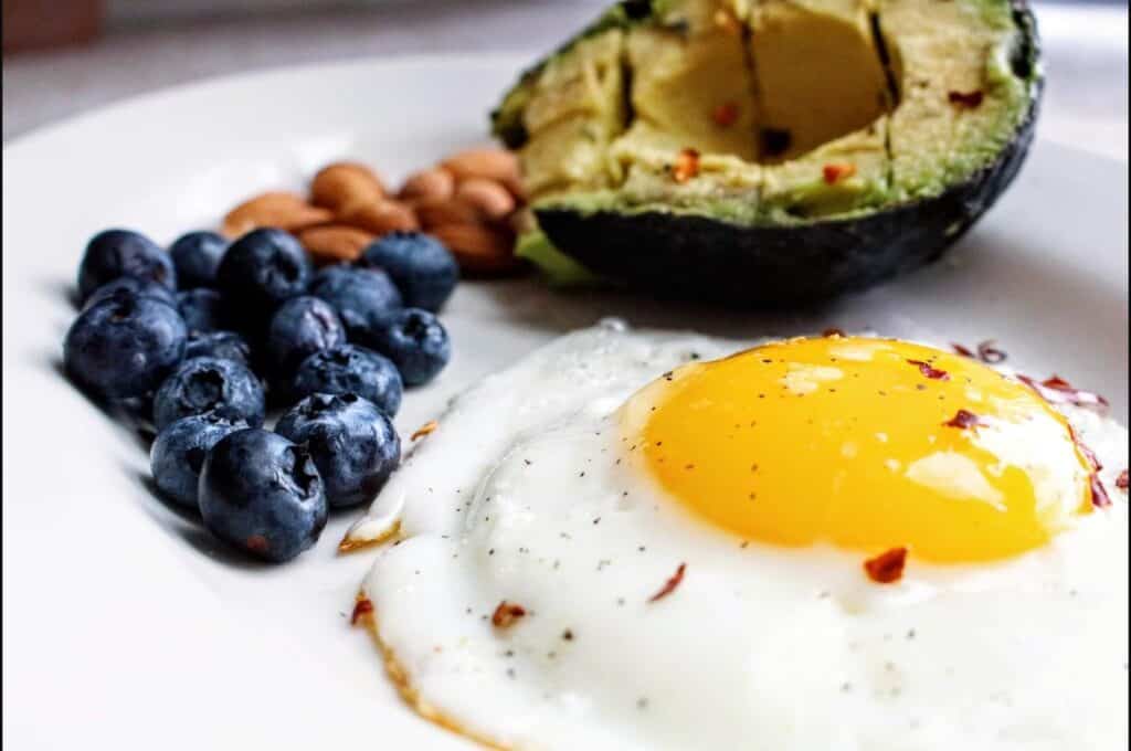 Plate with fried egg, avocado, blueberries and almonds representing protein, fat and carbohydrate food sources
