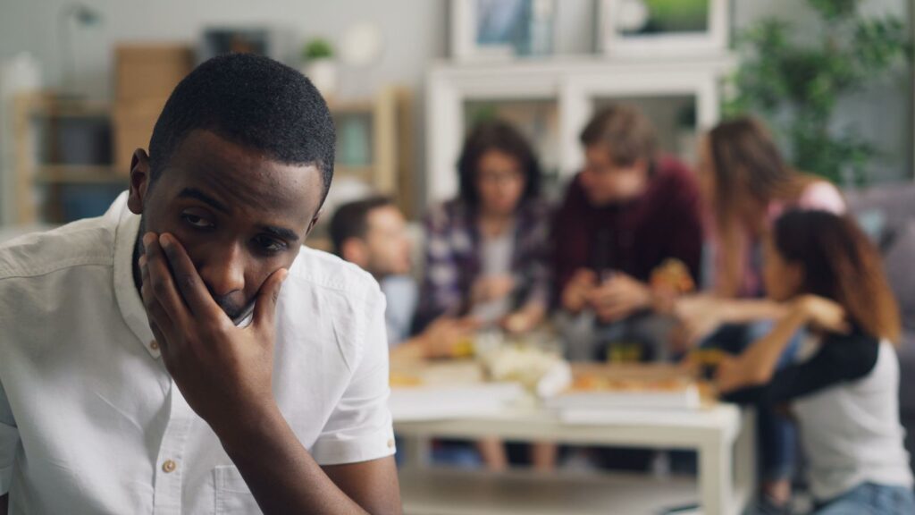 A concerned man sits apart from a lively group in a cozy home setting
