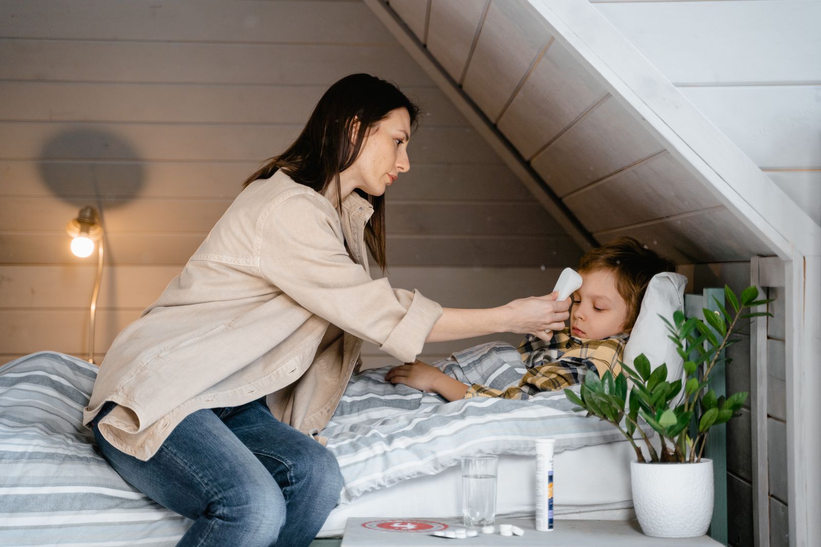 A mother checks her sick child's temperature in a cozy bedroom setting