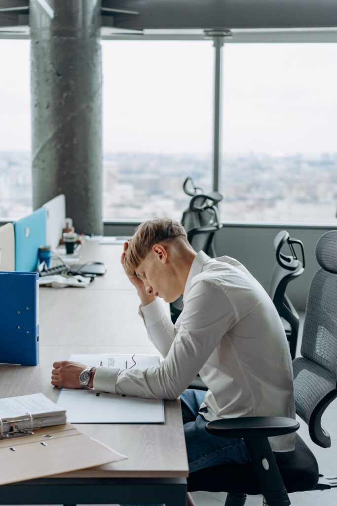 A stressed man sitting at a desk in a modern office, holding his head in his hands