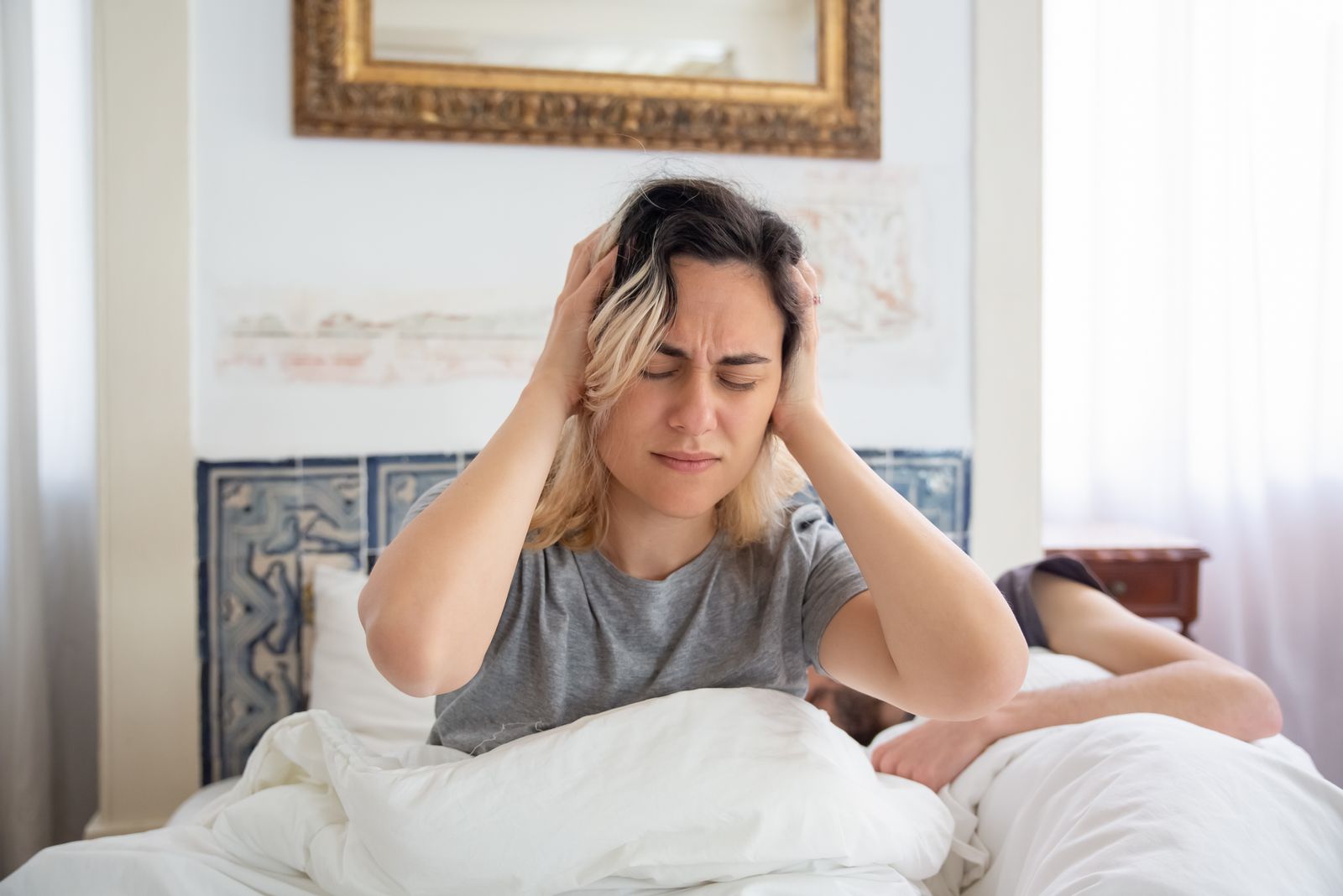 A woman holding her head in discomfort while sitting on a bed in the morning