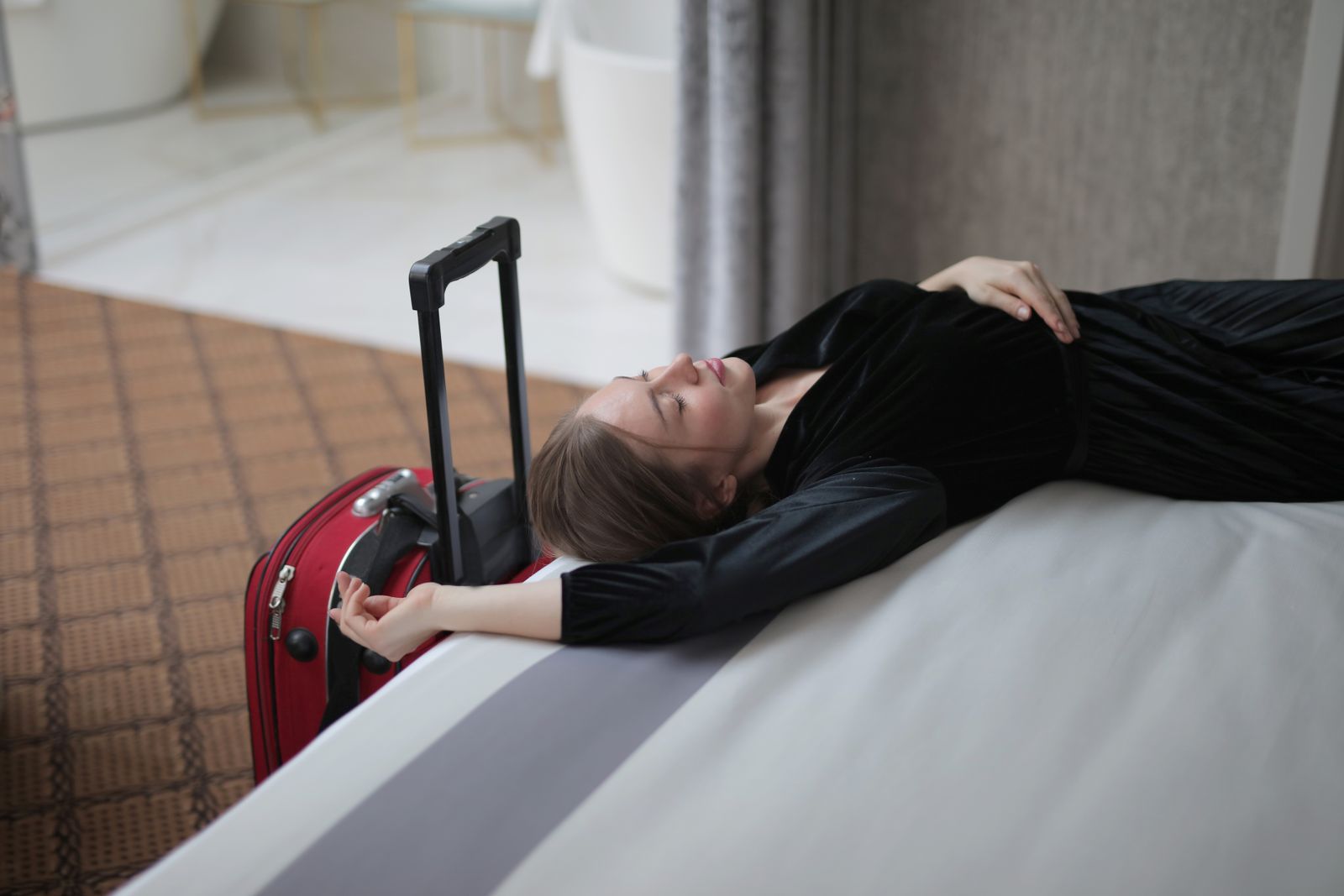 A woman resting on a hotel bed beside her suitcase