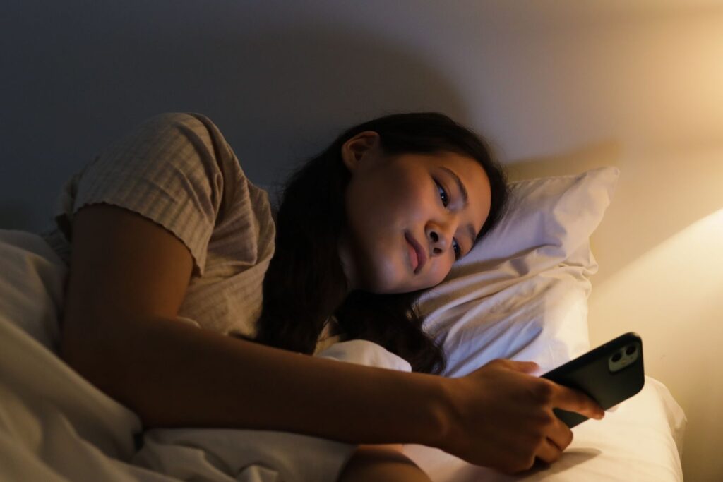 A woman lying in bed at night using a smartphone, illuminated by the screen light