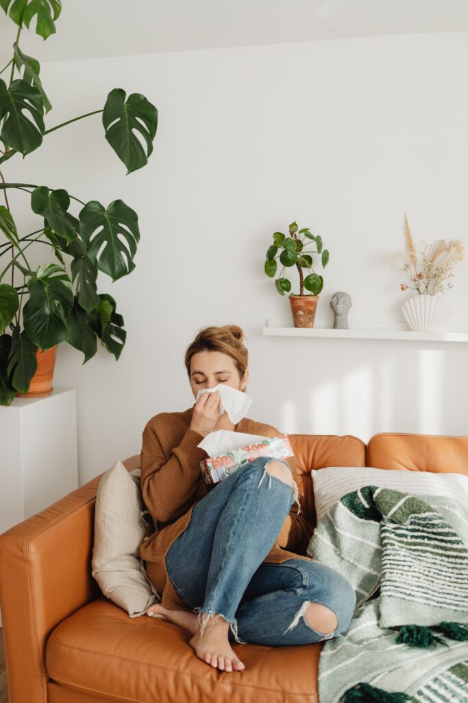 A woman sneezing into a tissue while sitting on a sofa surrounded by houseplants